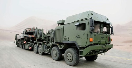 Rheinmetall MAN Military Vehicles HX81 tractor truck showing fully protected forward control cab and towing a semi-trailer carrying a Leopard 2 main battle tank with turret traversed to the rear.